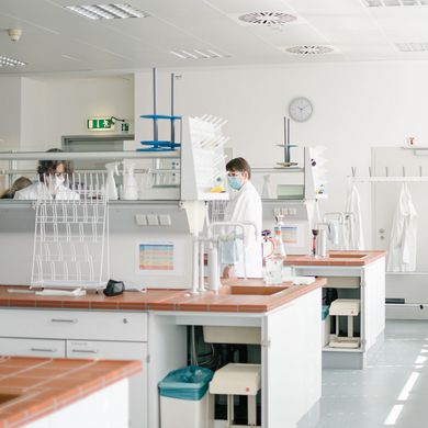 Im Hintergrund sind Studierende mit Masken in einem Labor zu sehen.