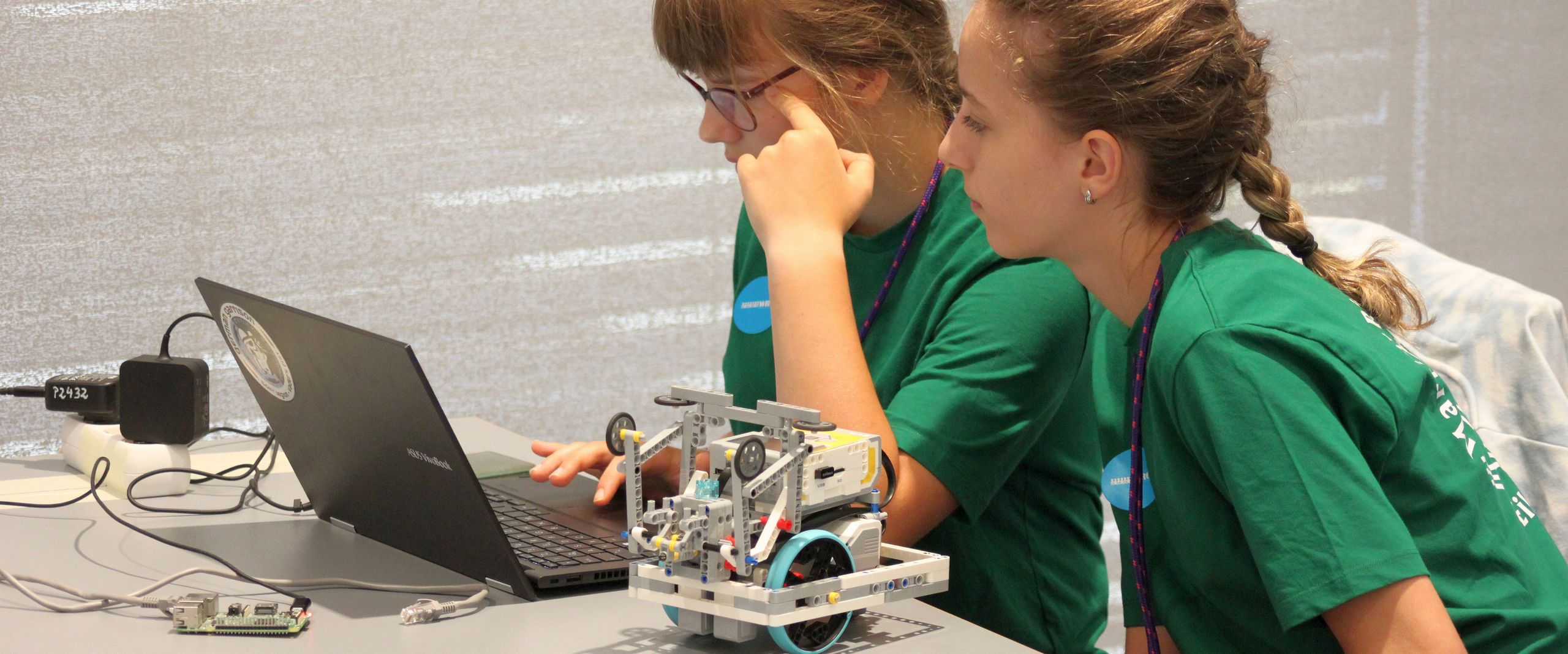 Zwei M&auml;dchen sitzen vor einem laptop und schauen auf dem Bildschirm. Auf dem Tisch neben ihnen steht ein kleiner Roboter auf R&auml;dern. Tisch und beobachten einen kleinen Roboter of R&auml;dern.