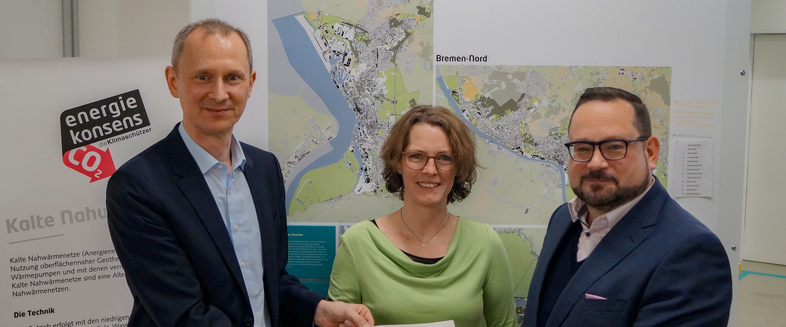 20240209 Foto DBU Förderbescheid Zwei Männer und eine Frau stehen vor einer Infotafel. Sie halten gemeinsam ein Papier und lächeln in die Kamera.