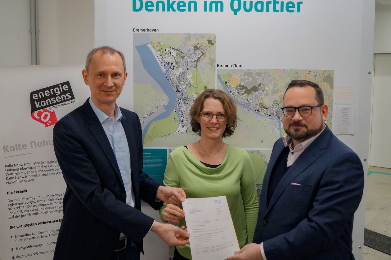 20240209 Foto DBU Förderbescheid Zwei Männer und eine Frau stehen vor einer Infotafel. Sie halten gemeinsam ein Papier und lächeln in die Kamera.