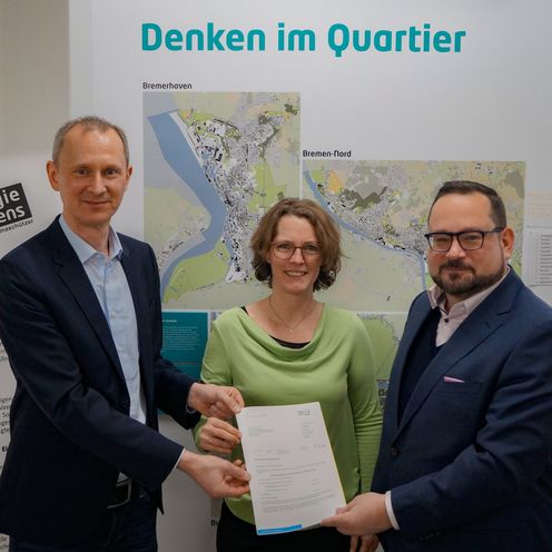 Zwei Männer und eine Frau stehen vor einer Infotafel. Sie halten gemeinsam ein Papier und lächeln in die Kamera.