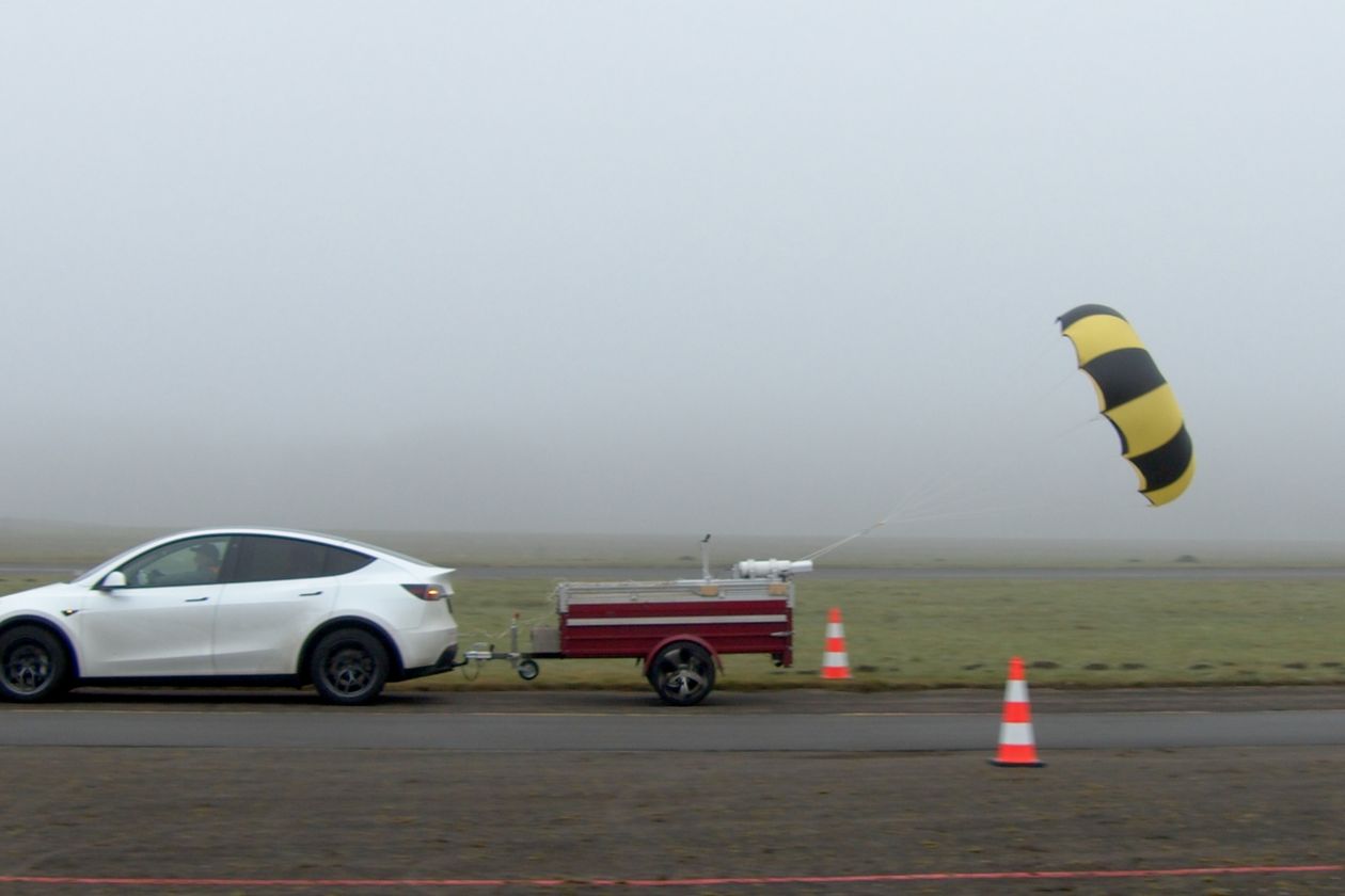 Das Bild zeigt ein fahrendes weißes Auto von der linken Seite welches einen kleinen roten Anhänger zieht. Auf dem Anhänger ist ein Testaufbau montiert, mit dem ein gelb-schwarzer geöffneter Fallschirm gezogen wird.