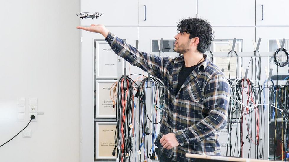 Auf dem Bild ist ein Student mit dunklen lockigen Haaren zu sehen. Er steht am Ende eine Vorlesungsraumes und streckt seine Hand nach oben aus. Kurz über der Hand fliegt eine Drohne. 