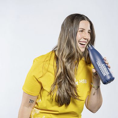 Lachende Studentin in einem gelben T-Shirt mit HSB-Logo und einer blauen Trinkflasche mit HSB-Logo in der Hand.