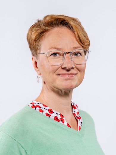 Auf dem Bild ist Claudia Schönherr zusehen. Sie hat kurzes rotblondes Haar. Sie trägt eine Brille und eine rot-weiße Bluse unter einem mintfarbenden Pullover. 