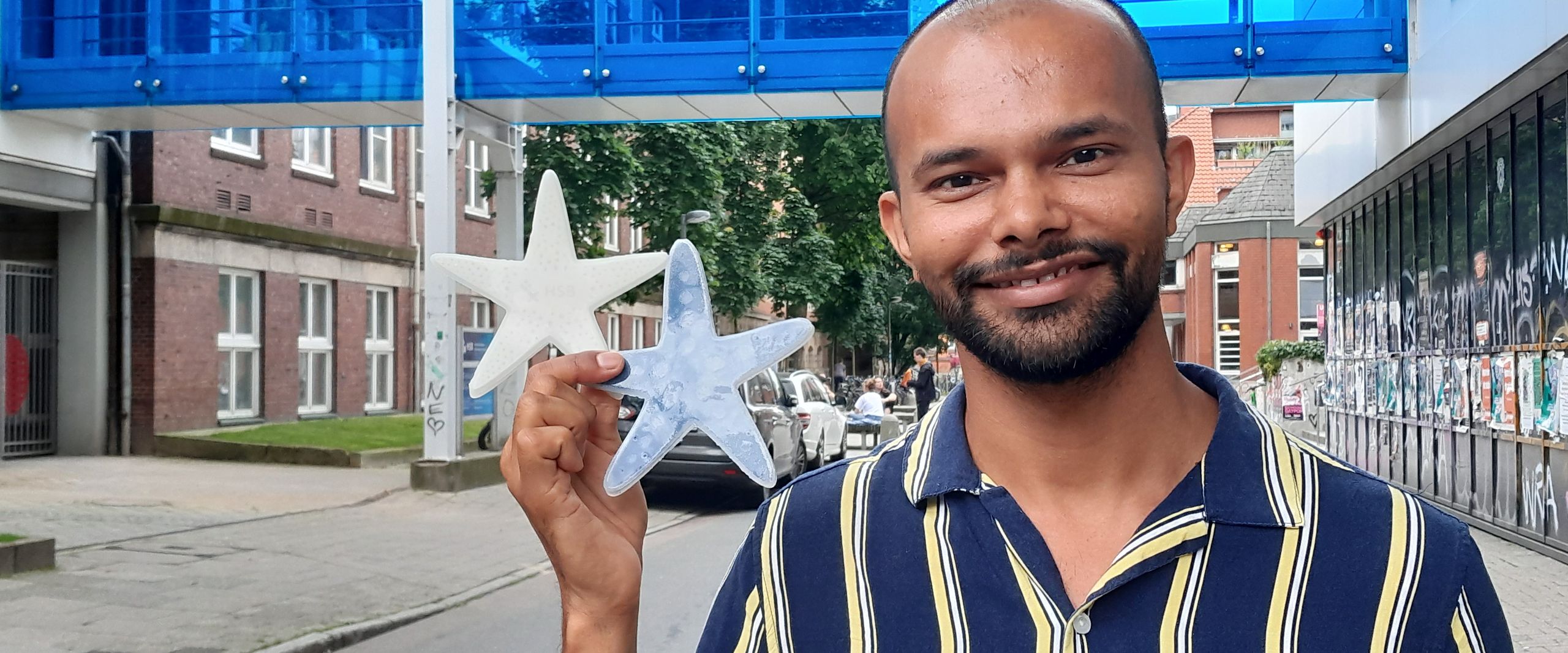 Raman Seestern Patent 2024 Portrait eines jungen mannes, der in der rechten hand zwei Seesterne aus Silikon hält: einer weiß, einer dunkel. Im Hintergrund sieht man eine blaue Fußgängerbrücke zwischen zwei Gebäuden.