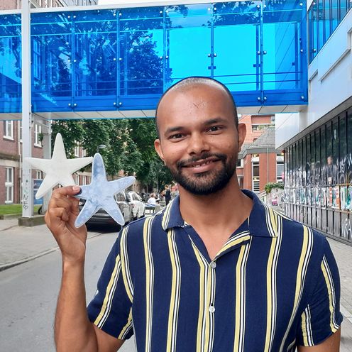 Portrait eines jungen mannes, der in der rechten hand zwei Seesterne aus Silikon h&auml;lt: einer wei&szlig;, einer dunkel. Im Hintergrund sieht man eine blaue Fu&szlig;g&auml;ngerbr&uuml;cke zwischen zwei Geb&auml;uden.