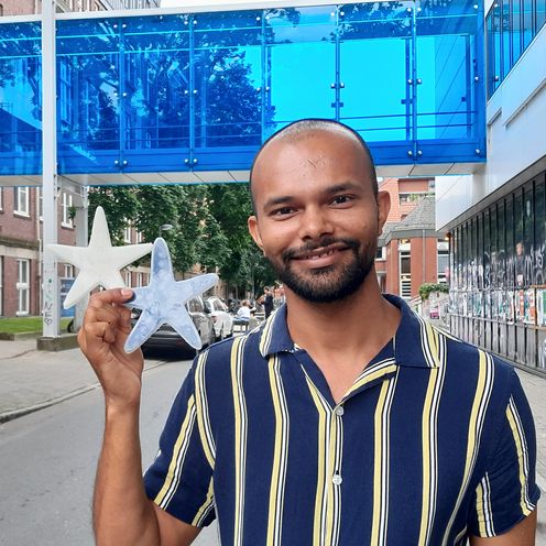 Portrait eines jungen mannes, der in der rechten hand zwei Seesterne aus Silikon h&auml;lt: einer wei&szlig;, einer dunkel. Im Hintergrund sieht man eine blaue Fu&szlig;g&auml;ngerbr&uuml;cke zwischen zwei Geb&auml;uden.