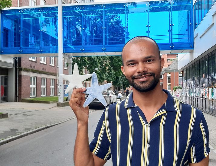 Portrait eines jungen mannes, der in der rechten hand zwei Seesterne aus Silikon hält: einer weiß, einer dunkel. Im Hintergrund sieht man eine blaue Fußgängerbrücke zwischen zwei Gebäuden.