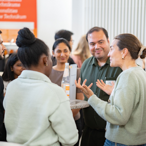 Menschen stehen um einen runden Stehtisch herum und unterhalten sich. Im Hintergrund sind weitere. Menschen zu sehen.