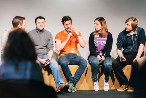 Fünf Studierende sitzen auf einer Bühne. Sie halten Mikrofone in der Hand. Ein Student, der in der Mitte spricht in das Mikrofon, die anderen schauen ihn interessiert an, einer lächelt in Richtung Publikum.