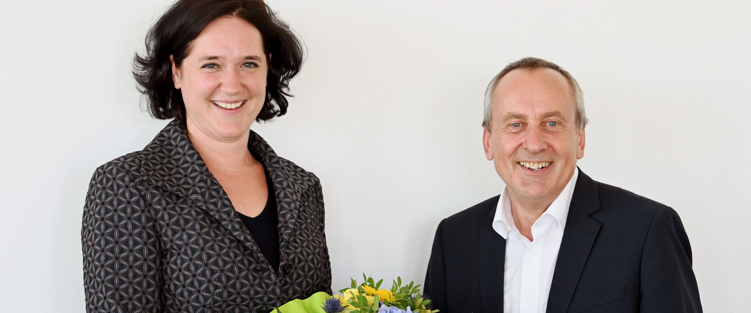 Frau und Mann lächeln in die Kamera. Sie halten einen Blumenstrauß und eine Urkunde.
