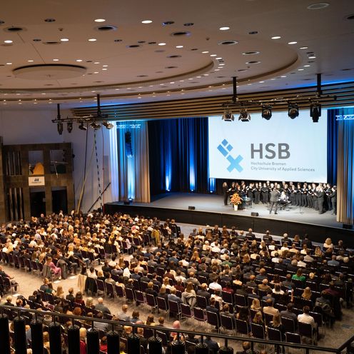 Menschen sitzen in einem Festsaal. Auf der B&uuml;hne steht ein Chor mit Dirigent und singt.