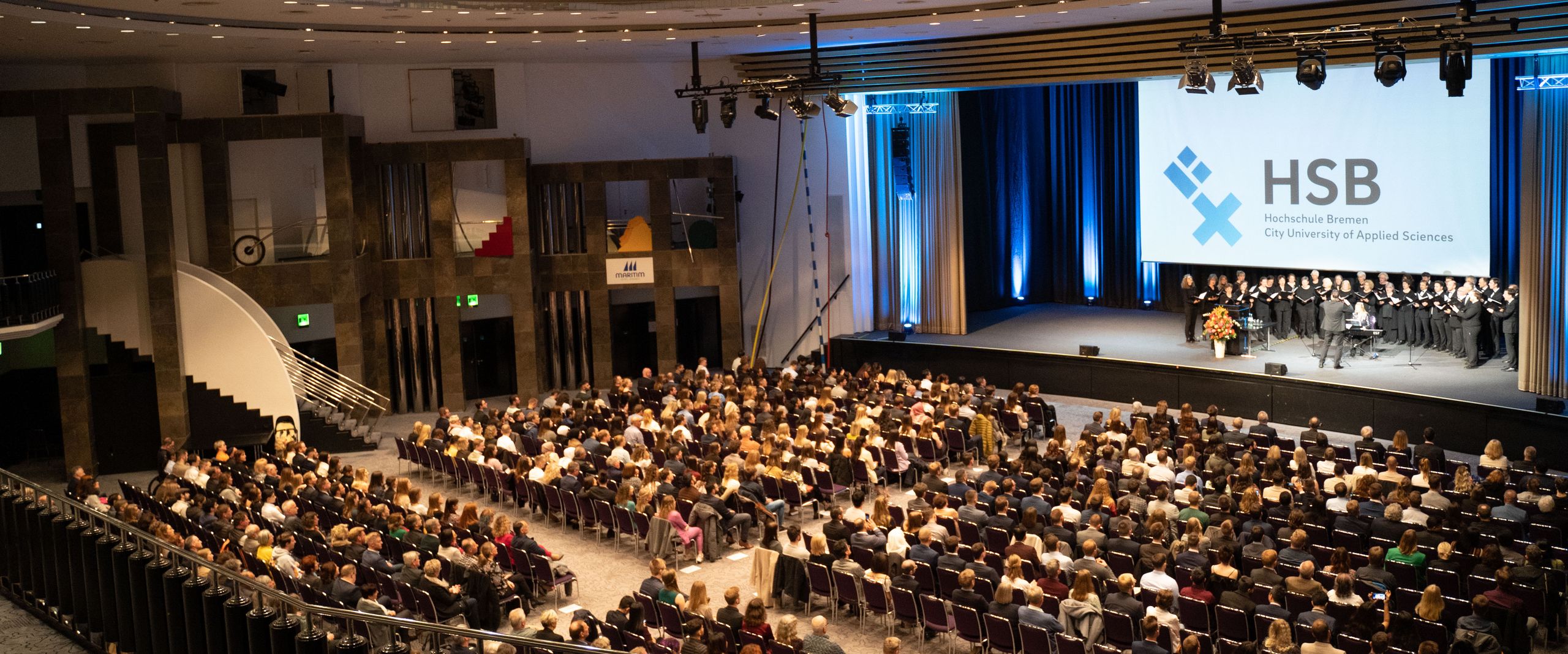 Menschen sitzen in einem Festsaal. Auf der Bühne steht ein Chor mit Dirigent und singt.