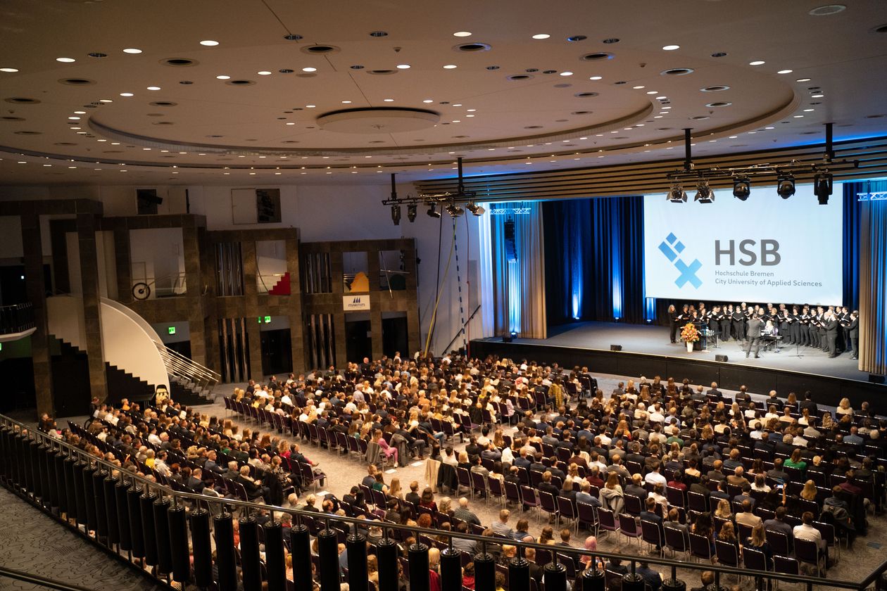 Menschen sitzen in einem Festsaal. Auf der Bühne steht ein Chor mit Dirigent und singt.