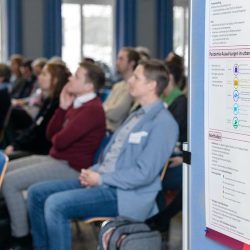 Zuh&ouml;rer:innen sitzen auf St&uuml;hlen. Im Vordergrund sieht man ein Banner mit Grafiken. 