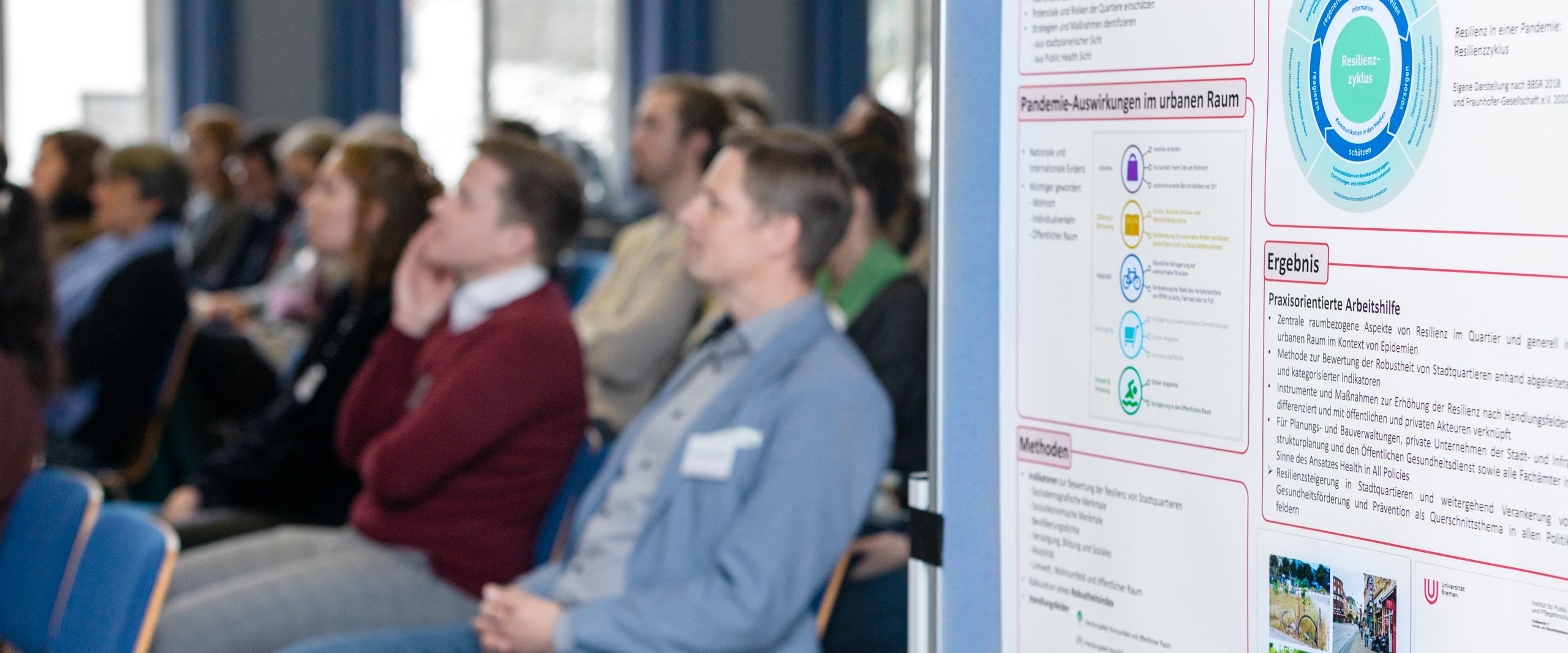 Zuh&ouml;rer:innen sitzen auf St&uuml;hlen. Im Vordergrund sieht man ein Banner mit Grafiken. 