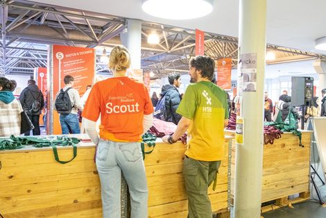 Zwei Studierende, in T-Shirts mit HSB-Logo gekleidet, stehen an einem Infostand mit HSB-Merchandise. Im Hintergrund sind Besucherinnen und Besucher des StudienINFOtags zu sehen, die im Gespräch sind oder Informationen auf Aufstellern ansehen.