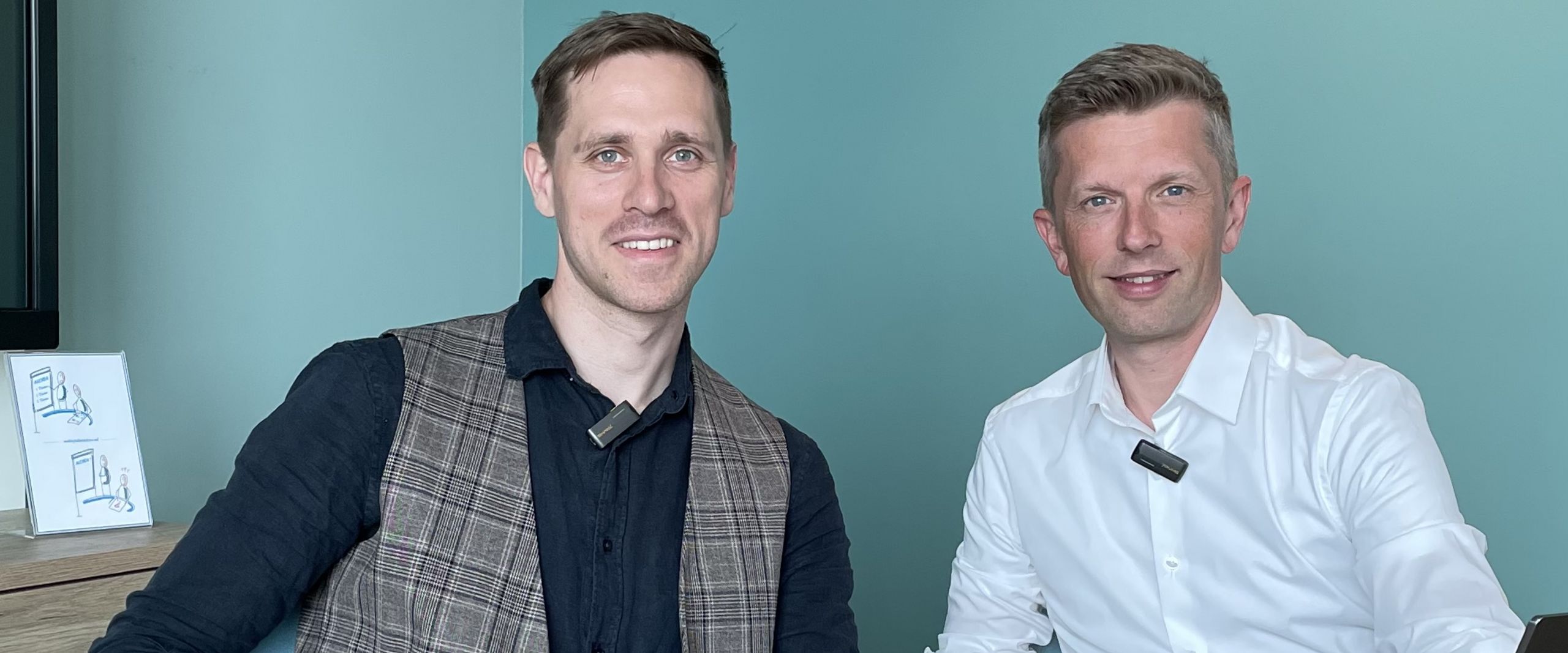 Dennis Kampen Sikora Dominik Uhr Zwei Männer sitzen am Tisch und alchen in die Kamera. Rechts von ihnen steht ein Laptop.
