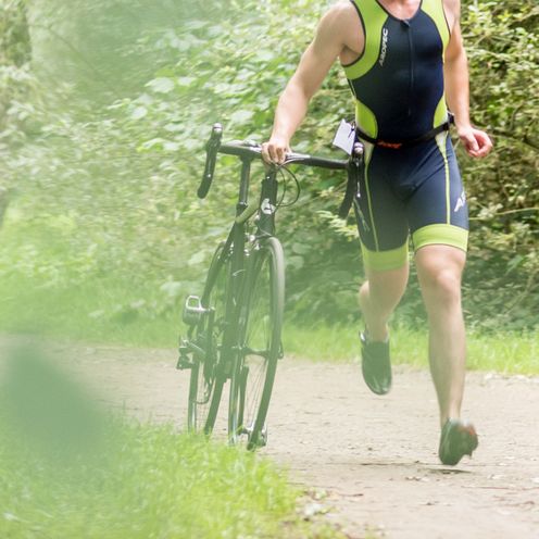 Ein Sportler l&auml;uft durch einen Wald und schiebt dabei ein Rennrad.
