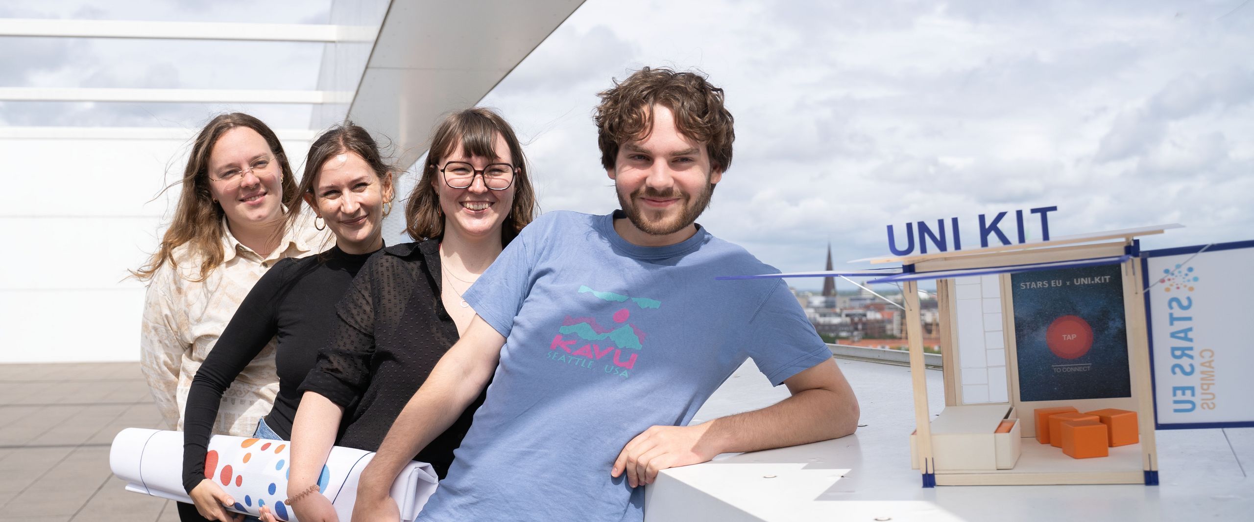 A young man and three young women stand one behind the other, laughing into the camera. Next to the man is a small wooden architectural model.