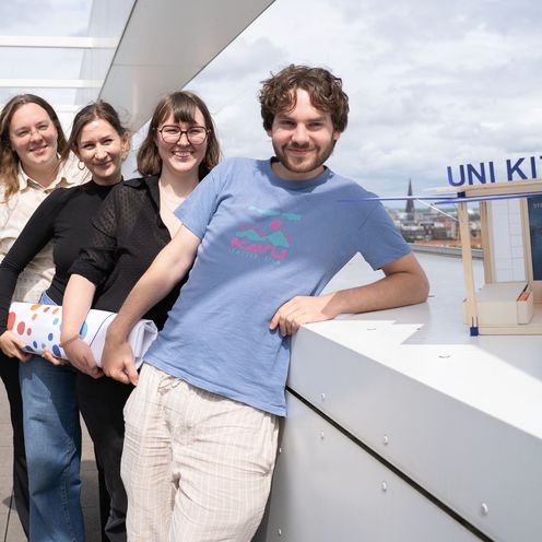 Interspace 2025 UniKit Ein junger Mann und drei junge Frauen stehen hintereinander und lachen in die Kamera. Neben dem Mann steht ein kleines Architekturmodell aus Holz.