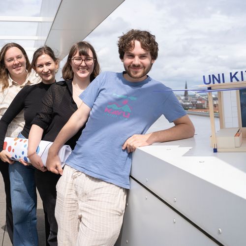 A young man and three young women stand one behind the other, laughing into the camera. Next to the man is a small wooden architectural model.