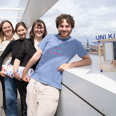 A young man and three young women stand one behind the other, laughing into the camera. Next to the man is a small wooden architectural model.