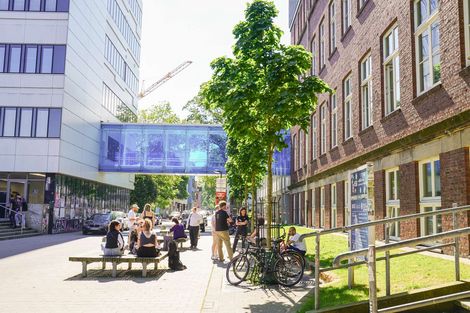 Studierende auf den Sitzgelegenheiten am Campus Neustadtswall bei sonnigem Wetter. Im Hintergrund sind das AB-Geb&auml;ude mit einer blauen verglasten Br&uuml;cke sowie das M-Geb&auml;ude zu sehen.