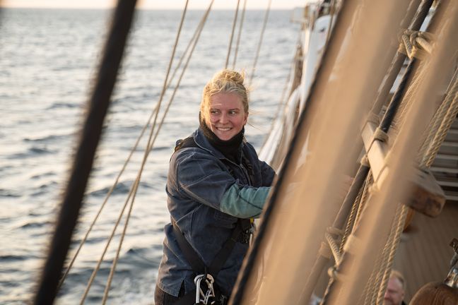 Auf dem Bild ist eine Frau zusehen, die in den Seilen eines Segelbootes klettert. 