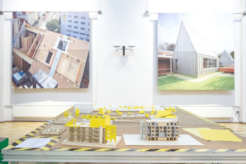 Das Bild zeigt Architekturmodelle auf einem Ausstellungstisch, im Hintergrund gro&szlig;formatige Fotografien von Bauprozessen und einem realisierten Geb&auml;ude.