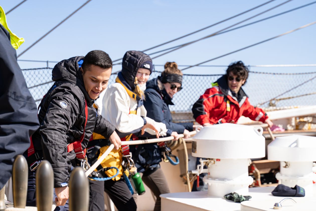 M&auml;nner und Frauen ziehen auf einem Segelschiff gemeisam an einem Tau.