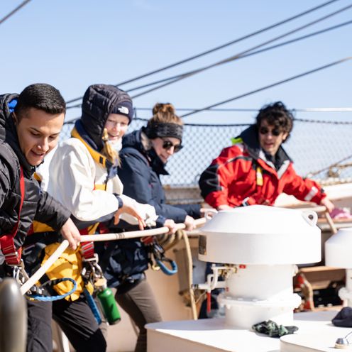 M&auml;nner und Frauen ziehen auf einem Segelschiff gemeisam an einem Tau.