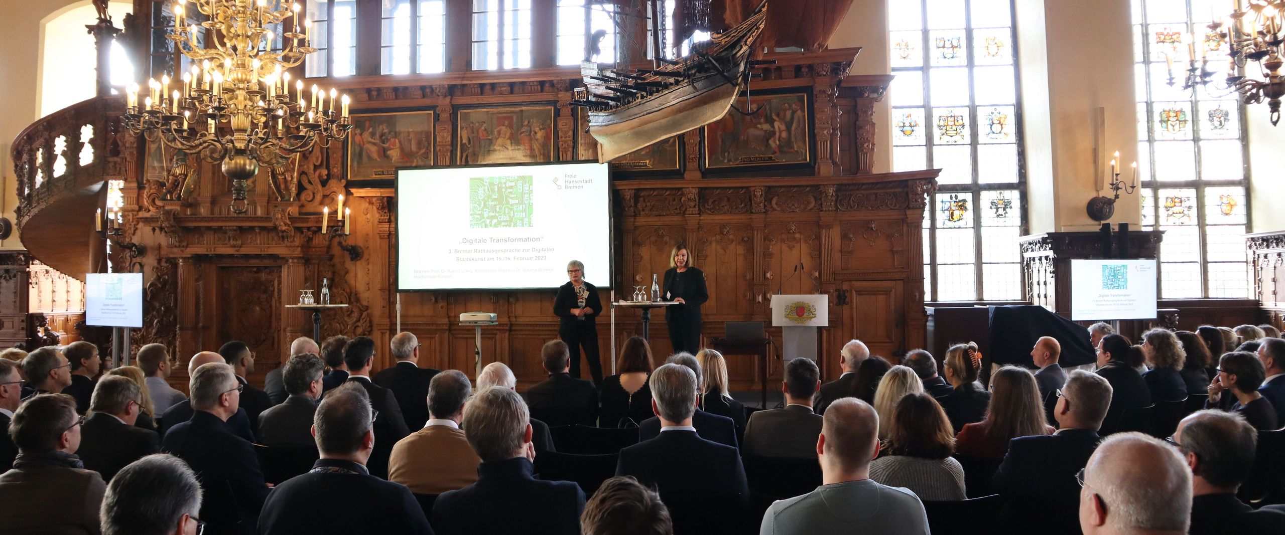 Menschen sitzen in einem großen Saal und hören zwei Rednerinnen zu. 