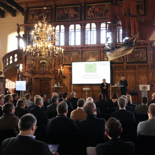 Menschen sitzen in einem gro&szlig;en Saal und h&ouml;ren zwei Rednerinnen zu. 