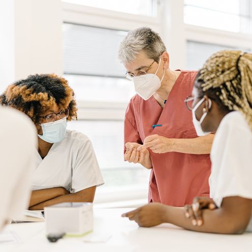 Menschen stehen an einem Tisch und besprechen sich. Sie haben zum Teil wei&szlig;e Kittel an.