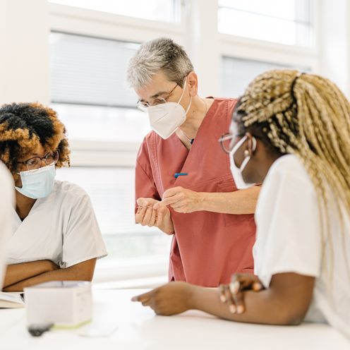 Menschen stehen an einem Tisch und besprechen sich. Sie haben zum Teil wei&szlig;e Kittel an.