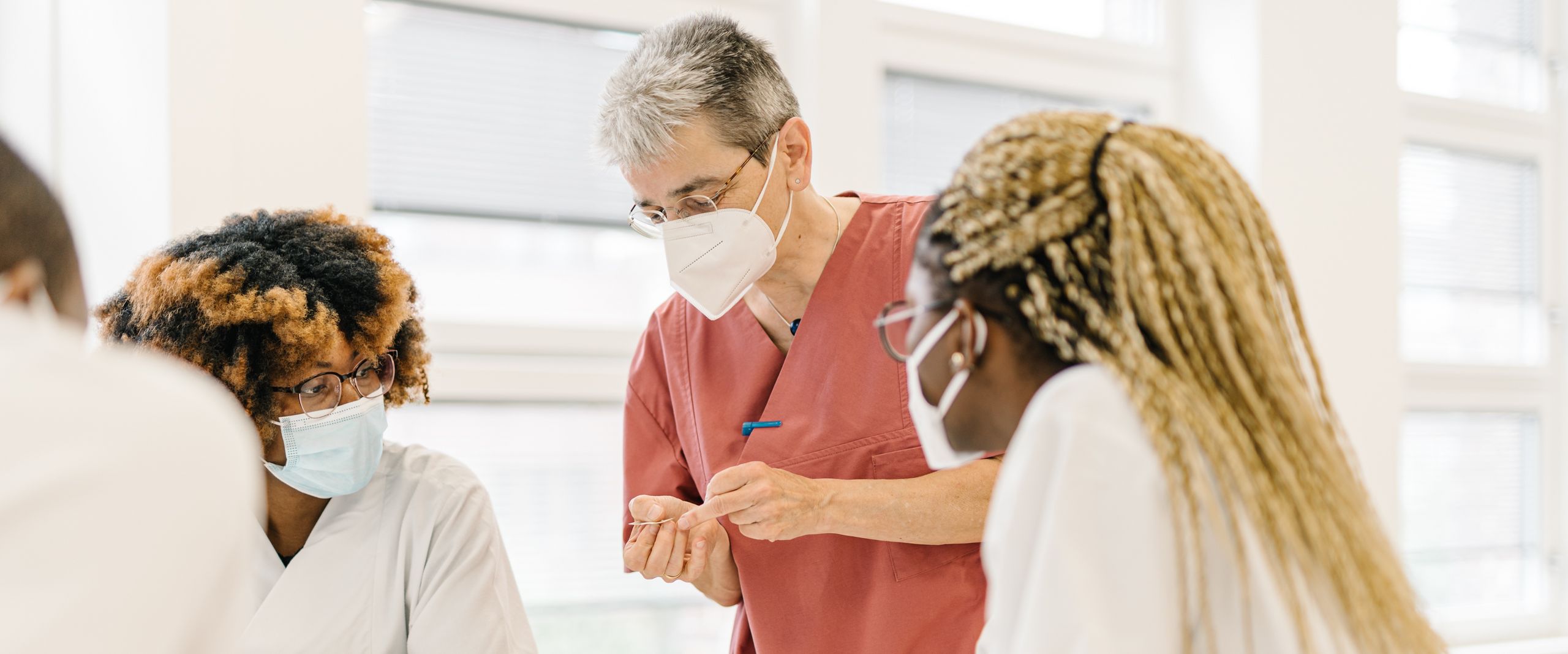 Menschen stehen an einem Tisch und besprechen sich. Sie haben zum Teil wei&szlig;e Kittel an.