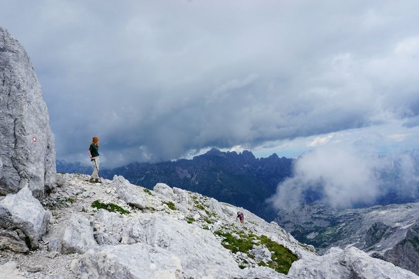Eine junge Frau steht an oben an einem steinigen Berghangs und schaut in die Ferne. Die Aussicht ist größtenteils von Wolken verhängt.