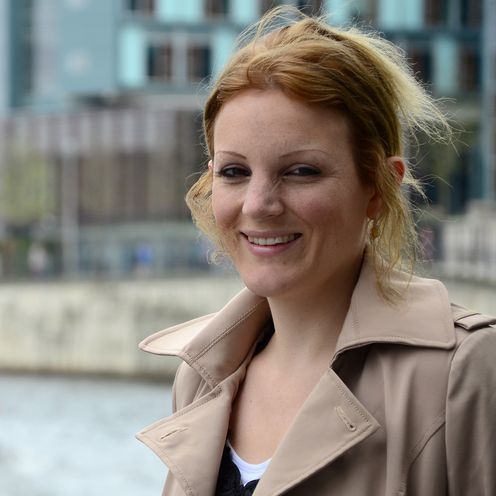 Frau stehend rechts im Bild mit l&auml;chelndem Blick in die Kamera. Im Hintergrund Wasser, darauf ein Boot. Au&szlig;erdem Geb&auml;ude.