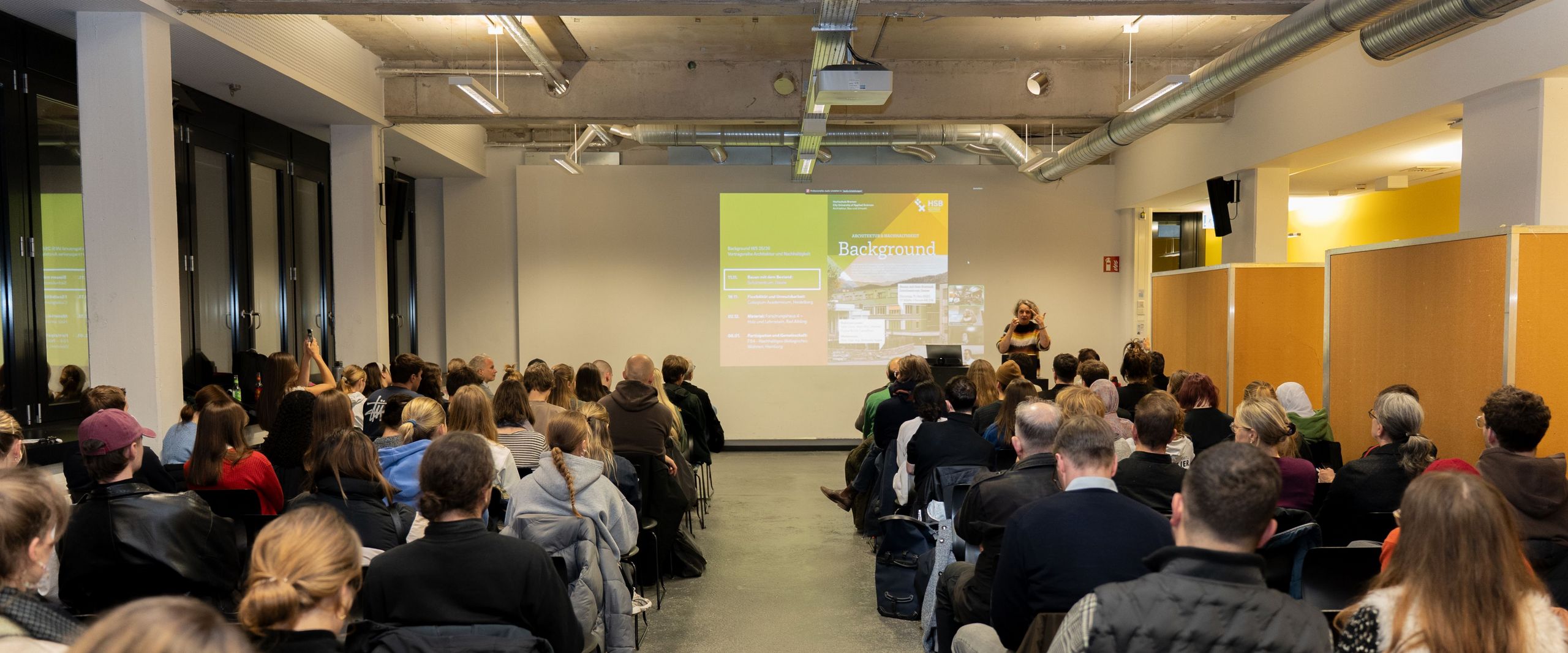 Gasvortragsreihe "Architektur und Nachhaltigkeit", Foto: Bettina Schürkamp (HSB) Publikum sitzt in einem modernen Veranstaltungsraum und hört einem Vortrag zu; vorne präsentiert eine Referentin eine Folie zum Thema Architektur und Nachhaltigkeit.