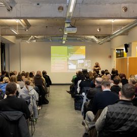 Publikum sitzt in einem modernen Veranstaltungsraum und h&ouml;rt einem Vortrag zu; vorne pr&auml;sentiert eine Referentin eine Folie zum Thema Architektur und Nachhaltigkeit.