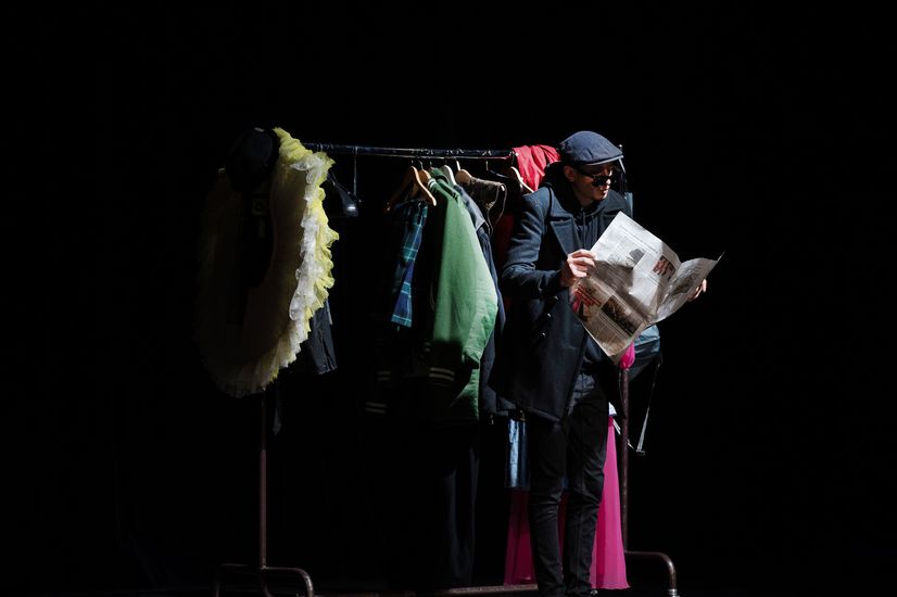 Mann mit Sonnenbrille steht mit Zeitung auf der B&uuml;hne vor einem Kleiderst&auml;nder