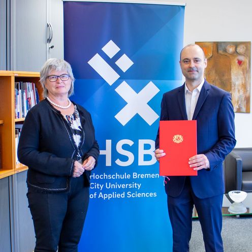 Frau und Mann stehen vor Banner. der Mann h&auml;lt eine Urkunde.