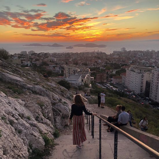 Fotografie, die im Hintergrund einen Sonnenuntergang über dem Meer vor Marseille zeigt. Im Vordergrund ist eine jungen Frau, die von hinten fotografiert eine Treppe hinuntergeht und dabei den Sonnenuntergang ansieht. un 