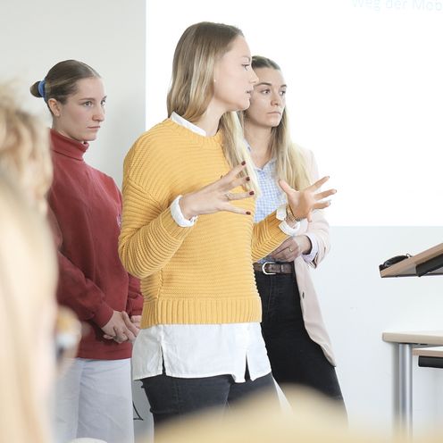 Auf dem Bild sind drei Studentinnen zu sehen, die vor einer Gruppe Menschen einen Vortrag h&auml;lt. In Hintergrund an einer wei&szlig;en Wand l&auml;sst sich eine Pr&auml;sentation erahnen. 