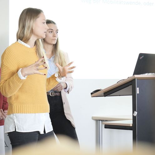 Auf dem Bild sind drei Studentinnen zu sehen, die vor einer Gruppe Menschen einen Vortrag h&auml;lt. In Hintergrund an einer wei&szlig;en Wand l&auml;sst sich eine Pr&auml;sentation erahnen. 