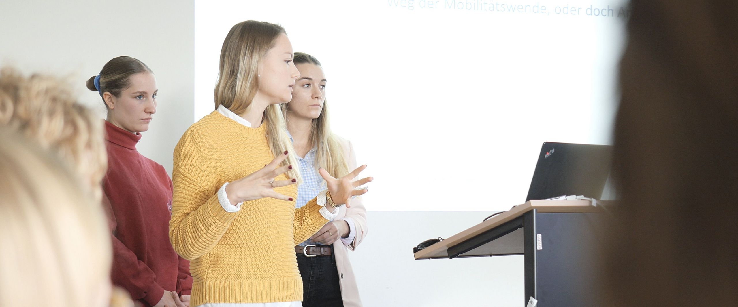 Auf dem Bild sind drei Studentinnen zu sehen, die vor einer Gruppe Menschen einen Vortrag hält. In Hintergrund an einer weißen Wand lässt sich eine Präsentation erahnen. 