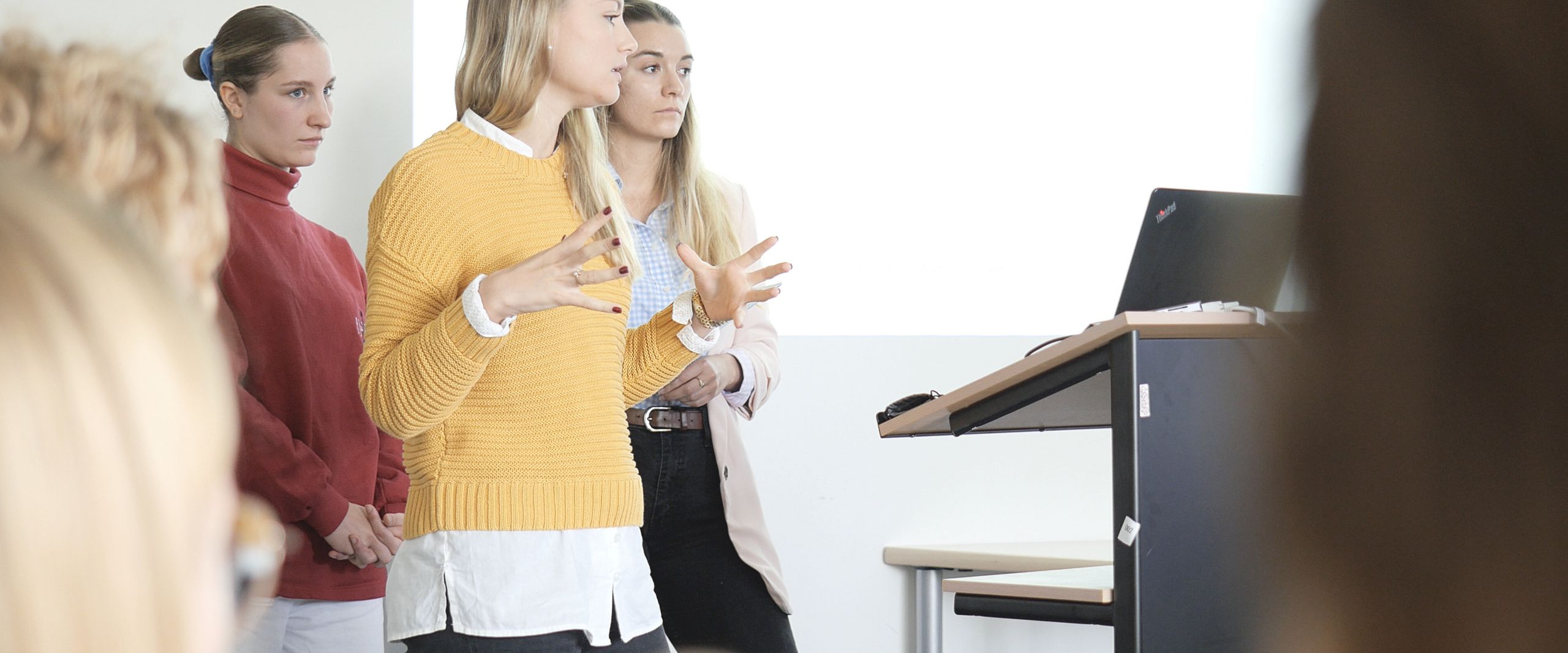 Vortragssituation INA Auf dem Bild sind drei Studentinnen zu sehen, die vor einer Gruppe Menschen einen Vortrag hält. In Hintergrund an einer weißen Wand lässt sich eine Präsentation erahnen.