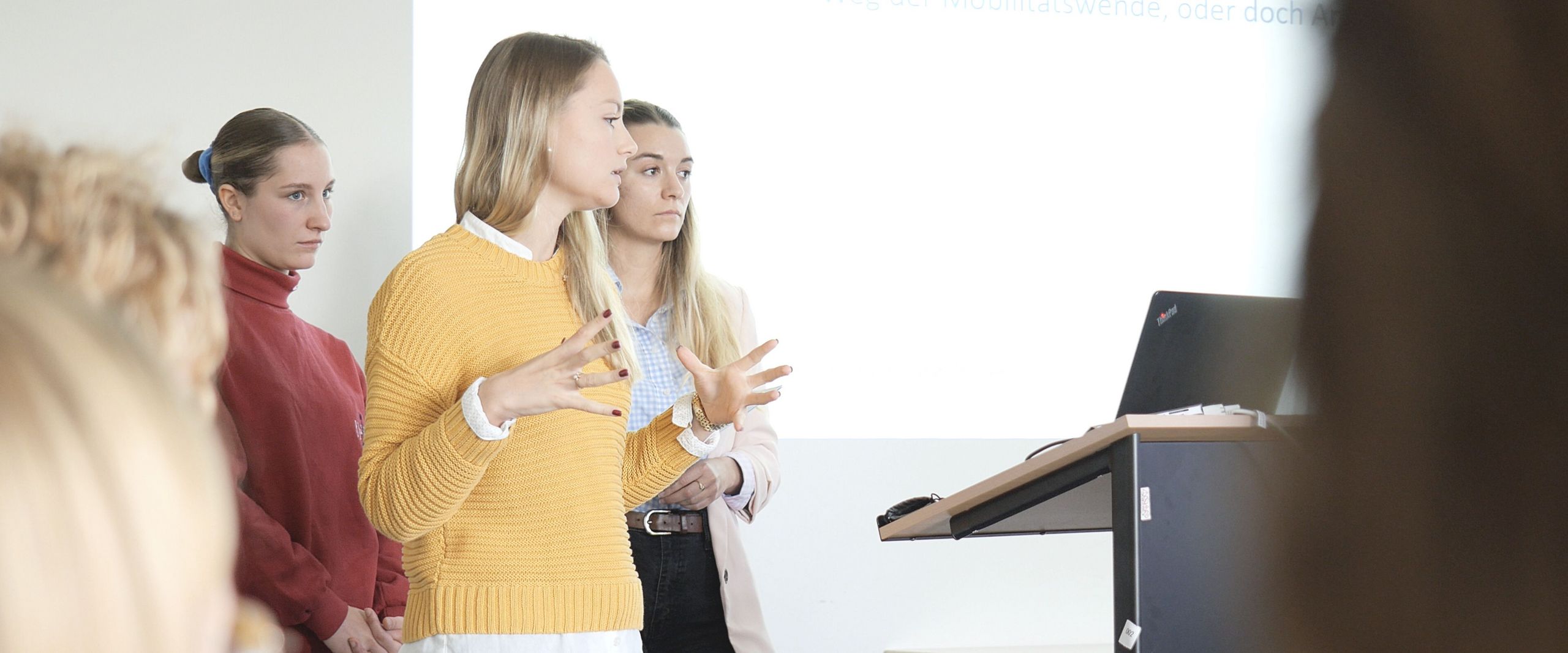 Auf dem Bild sind drei Studentinnen zu sehen, die vor einer Gruppe Menschen einen Vortrag hält. In Hintergrund an einer weißen Wand lässt sich eine Präsentation erahnen. 